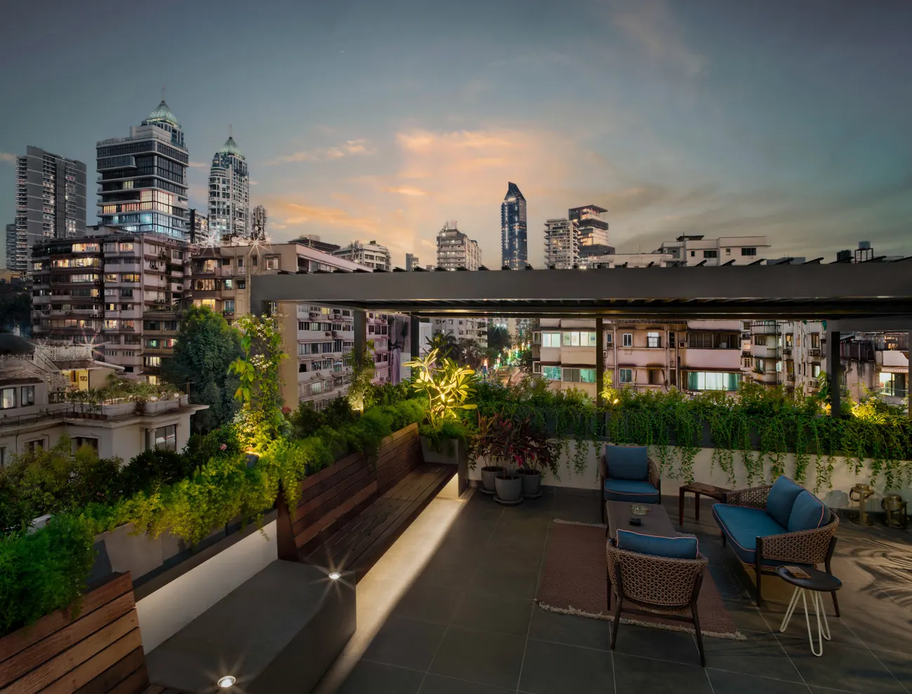 Rooftop terrace at Cambridge Court at dusk, with a louvred pergola, timber benches and planting against the Mumbai skyline.
