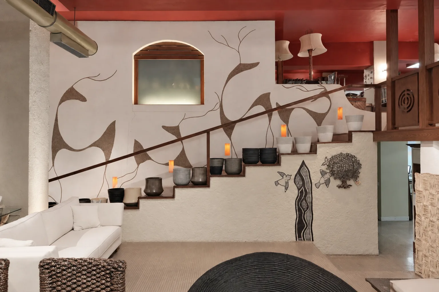 Stair wall at the Abaca showroom in Mumbai: a white plastered wall hand-drawn with gazelle figures in textured earth tones, above a stair lined with grey and white planters and small warm lamps, under a red ceiling.