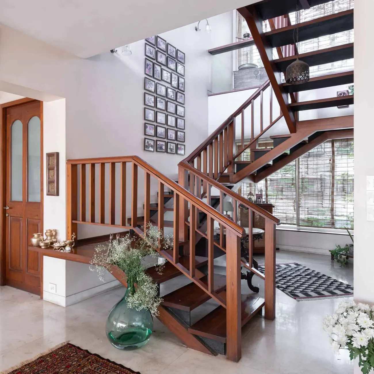 Double-height stair hall at Lloyds Gardens: a teak staircase with open treads rising past a wall of framed photographs toward bamboo-screened windows.
