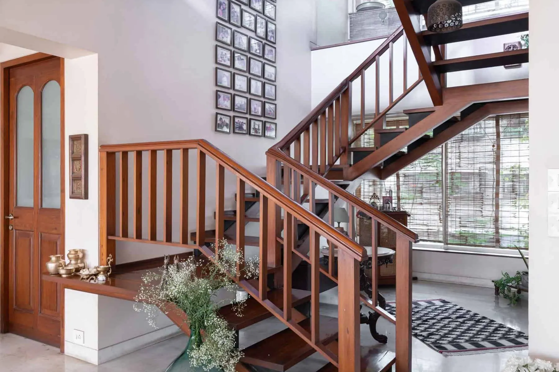 Double-height stair hall at Lloyds Gardens: a teak staircase with open treads rising past a wall of framed family photographs toward bamboo-screened windows.