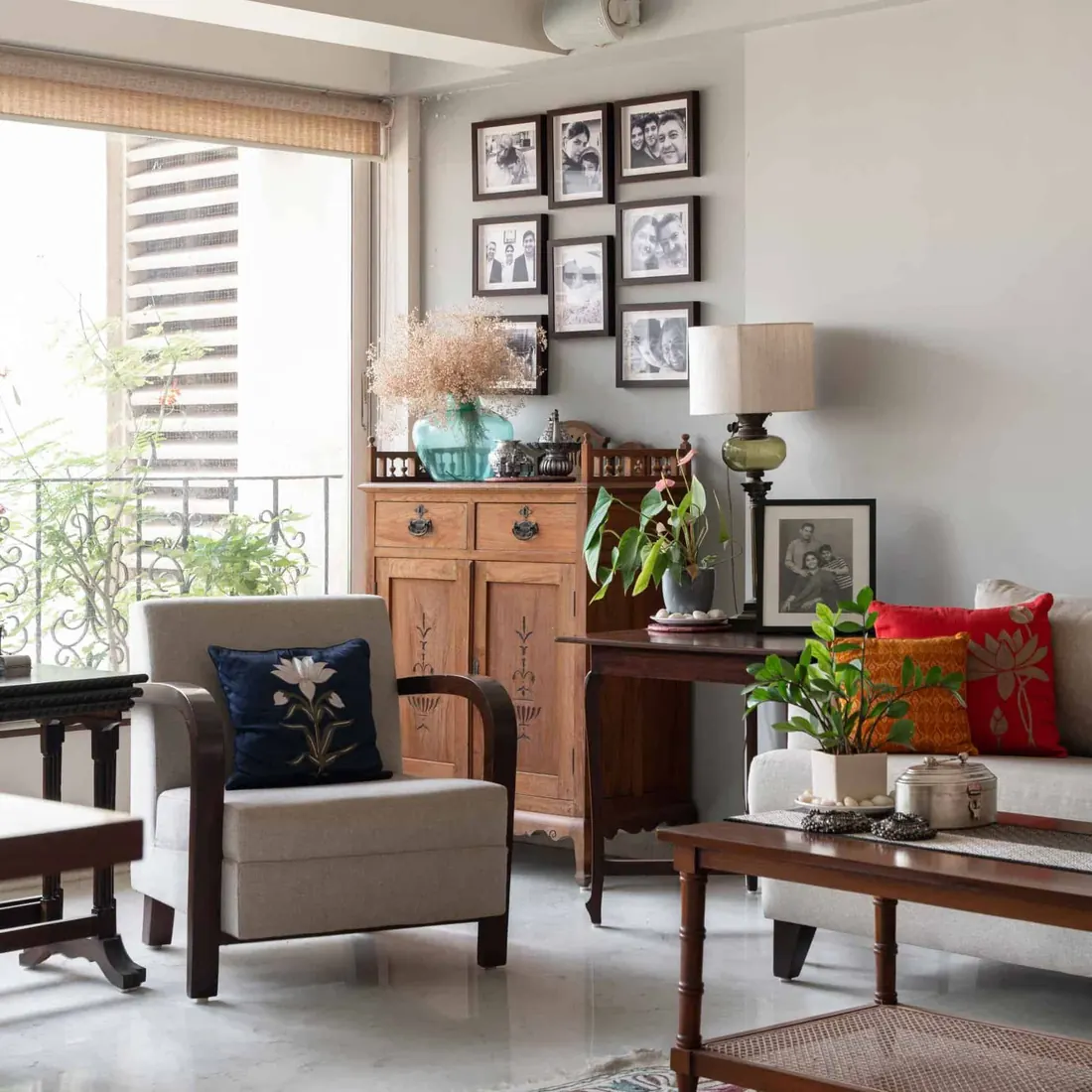 Living room corner at Lloyds Gardens with a linen armchair, an antique teak cabinet, a grid of black-and-white family photographs and a bamboo blind over the window.