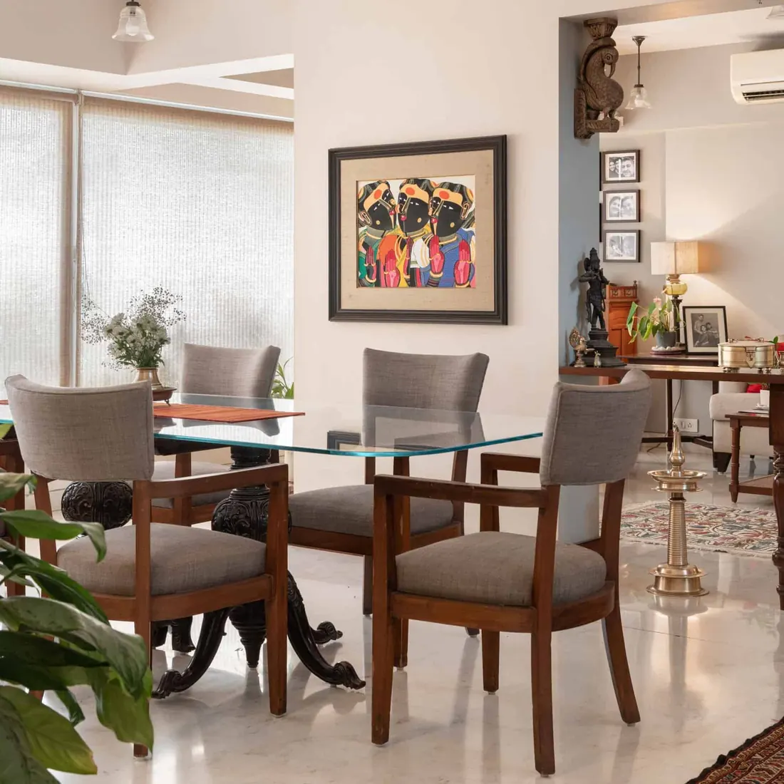 Dining area at Lloyds Gardens with a glass-topped table on a carved pedestal base, grey upholstered chairs, a framed painting and a brass lamp.