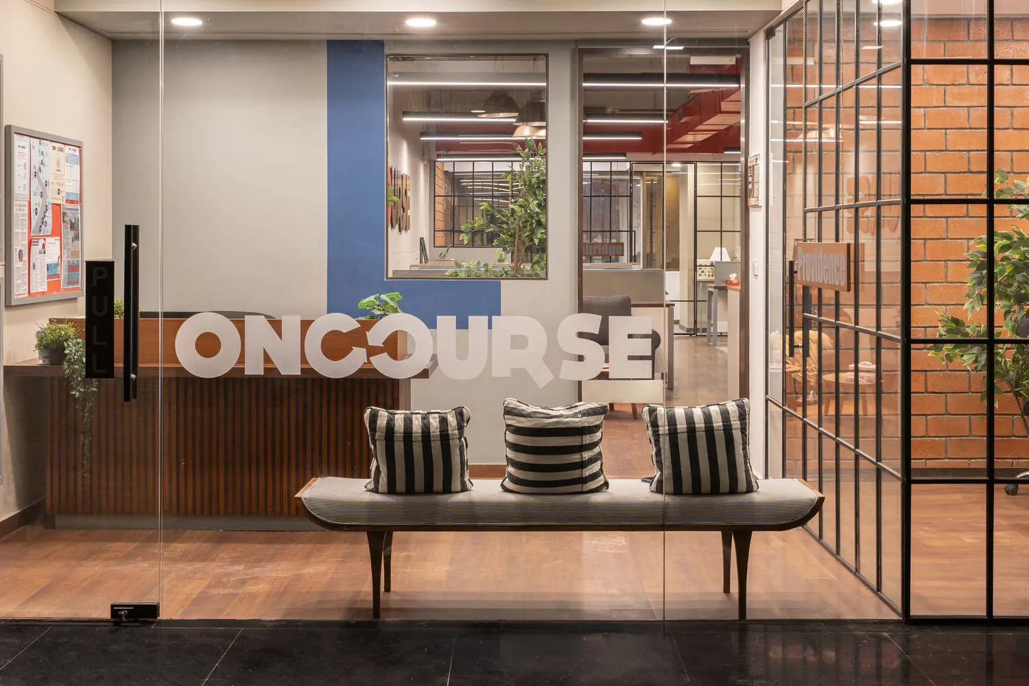 Glass entrance door at OnCourse etched with the OnCourse wordmark, with a striped bench and brick wall beyond.