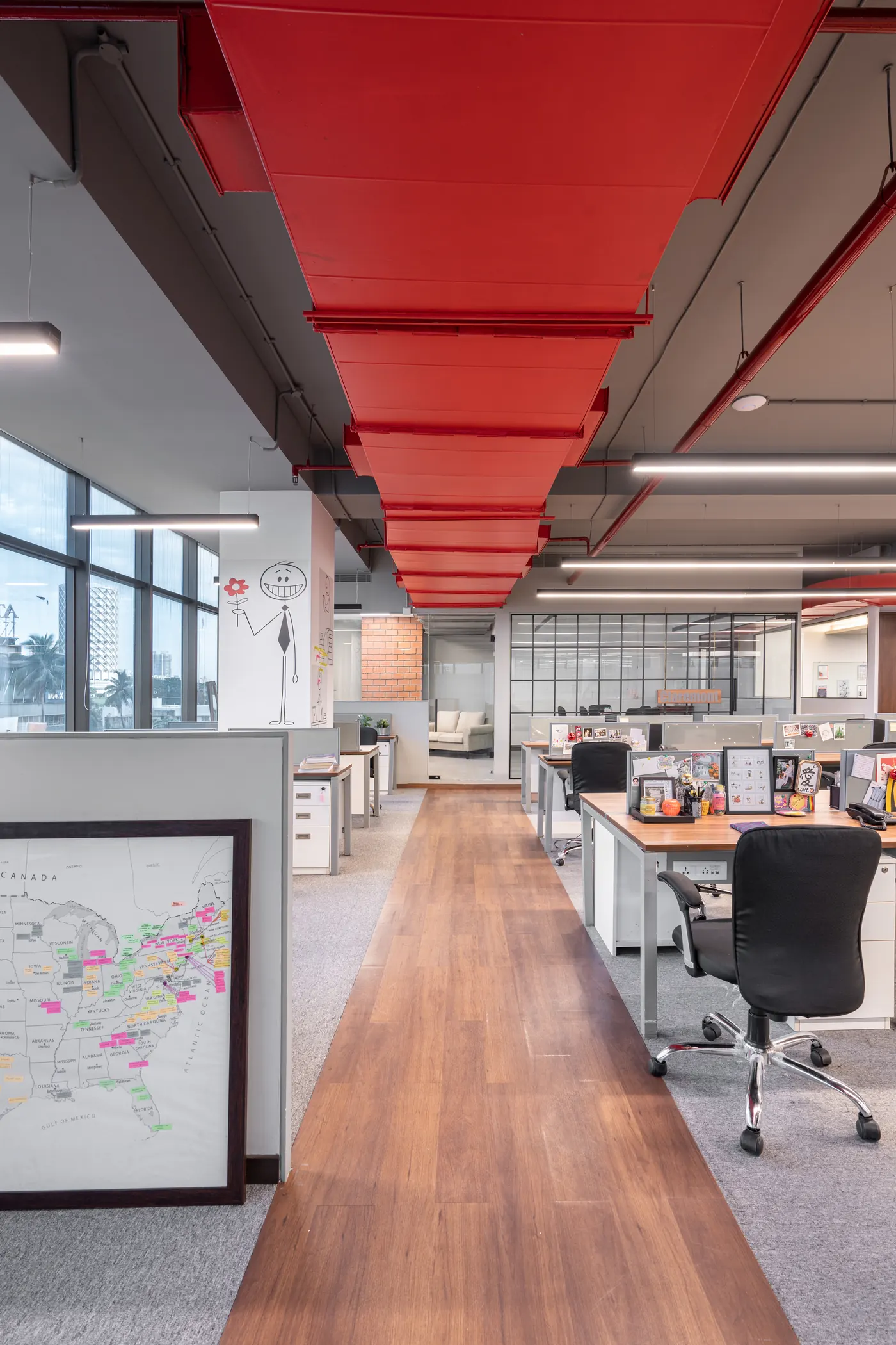 Open-plan OnCourse office in Worli: a red duct runs above a timber walkway between desks, with a framed map in the foreground.