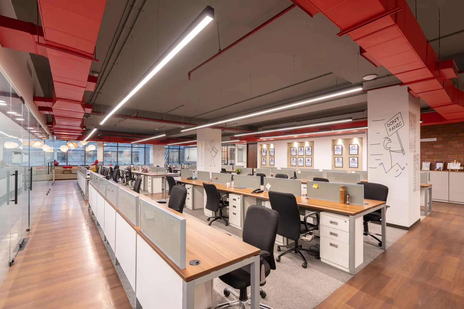 Workstations at OnCourse in Worli under red ducts and linear lights, with a column drawn with a line figure and a wall of framed student profiles beyond.