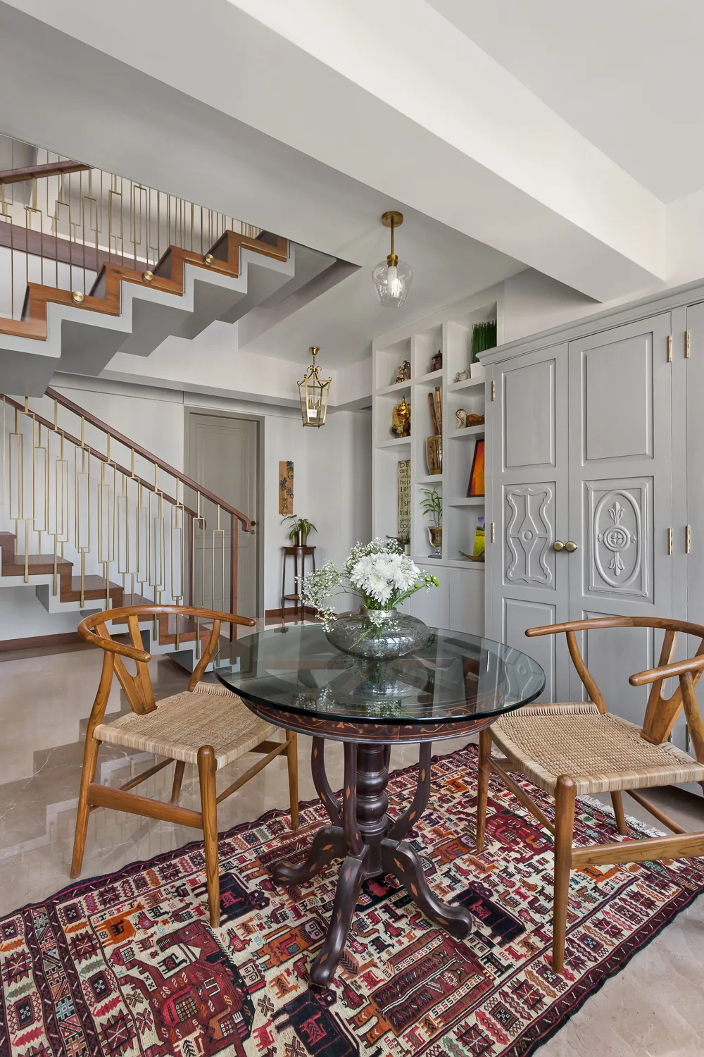 Breakfast table at Zara with wishbone chairs and an inlaid pedestal table on a kilim, beneath a staircase with a geometric brass railing, beside carved grey cupboard doors.