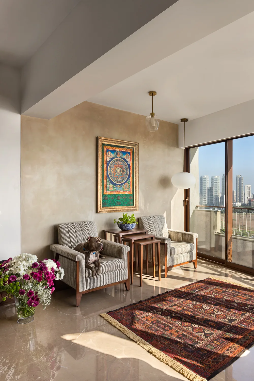 Sitting area at Zara: a framed thangka on a lime-plastered wall, striped armchairs with a dog resting on one, nesting tables and a kilim, with the city skyline beyond.